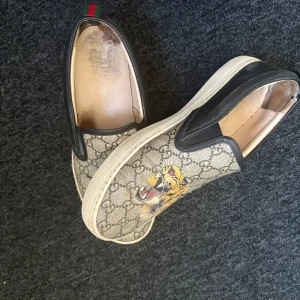 Gucci slip-on sneakers med tigertryck - Snygga slip-on sneakers från Gucci med klassiskt GG-mönster och färgglatt tigertryck på ovandelen. Skorna har svart läderdetalj vid hälen och vit platt sula. Perfekta för dig som vill sticka ut med exklusiv streetstyle.