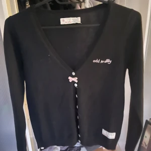 Svart kofta från Odd Molly med rosett i stork 0 - Säljer en svart kofta från Odd Molly med v-ringning och knappar framtill. Detalj med liten rosa rosett vid knäppningen och broderad logga på bröstet. Långärmad och mjuk i materialet, perfekt för lager-på-lager. Denna kofta är knappt använd och kostar cirka 1500kr i nypris.