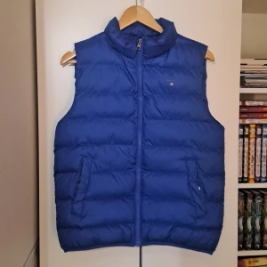 Blå dunväst från GANT - Snygg blå dunväst från GANT med dragkedja framtill och två fickor på sidorna. Västen har en hög krage och diskret GANT-logga på bröstet. Perfekt för lager-på-lager och håller dig varm under kyliga dagar. Kan förhandla vid snabbt köp. Passar även 180>