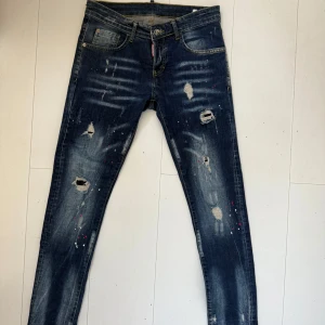 Mörkblå slitna jeans från Dsquared2 - Säljer ett par mörkblå jeans från Dsquared2 med slitningar, hål och färgstänk. Klassisk femficksmodell med smal passform. Perfekta för sommaren