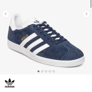 Adidas Gazelle mörkblå sneakers - Säljer ett par klassiska Adidas Gazelle sneakers i mörkblå mocka med vita detaljer och de ikoniska tre ränderna på sidan. Skorna har snörning och vit sula, samt guldfärgad Gazelle-text på sidan. Perfekta för en stilren och sportig look.