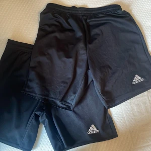 Svarta träningsshorts från Adidas - Två par svarta träningsshorts från Adidas med klassisk logga på ena benet. De har elastisk midja och är tillverkade i ett lätt och syntetiskt material som passar perfekt för sport och aktivitet.
