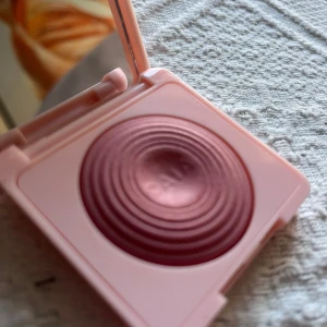 CAIA Glow Blush Raspberry Passion - En snygg rouge från CAIA i nyansen Raspberry Passion. Kommer i en ljusrosa, fyrkantig dosa med spegel. Rougen har en skimrande, bärig rosa ton och en vågig, upphöjd yta. Perfekt för att ge kinderna en fräsch glow.