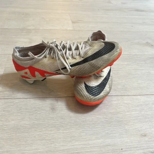 Nike Mercurial fotbollsskor i beige och röd original pris 1750 - Säljer ett par Nike Mercurial fotbollsskor i beige med svarta och röda detaljer. Skorna har snörning, rund tå och platt sula med dobbar för bra grepp på planen. Perfekta för dig som vill ha lätta och snabba skor till fotbollsträningen.