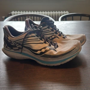 Vita och svarta löparskor från Saucony - Säljer ett par Saucony löparskor i vitt med svarta och grå detaljer samt blå rand på sulan. Skorna har snörning och mönstrad häl. Perfekta för träning och löpning, med lätt och luftig design.