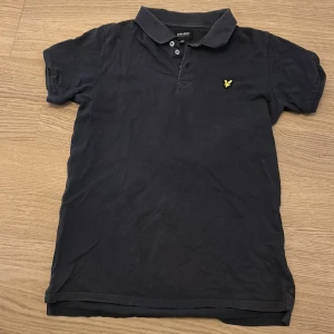 Mörkblå pikétröja från Lyle & Scott Junior - Säljer en mörkblå pikétröja från Lyle & Scott Junior med klassisk krage och knappar framtill. Tröjan har det ikoniska gula fågel-loggan broderad på bröstet. Perfekt för en stilren och avslappnad look.