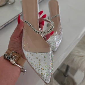 Glittriga klackskor med strassdetaljer - Säljer ett par transparenta klackskor med spetsig tå och glittriga stenar över hela skon. Skorna har en elegant rem med strass och en unik genomskinlig klack. Perfekta för dig som vill sticka ut med extra bling.