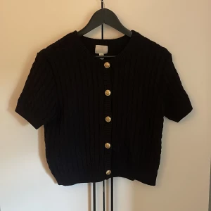 Svart stickad kofta med kort ärm från H&M - Kortärmad svart stickad kofta från H&M med ribbad struktur och guldknappar framtill.  Klassisk rund halsringning och kortare passform. Något liten i storlek 