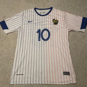 Frankrike Fotbollströja - Säljer en vit Frankrike fotbollströja med blå och röda ränder, nummer 10 och Mbappé på ryggen. Tröjan har blå detaljer vid krage och ärmslut samt broderad tupp-logga på bröstet. Tillverkad i Dri-Fit material för bra komfort.