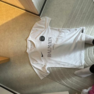 Vit PSG x Balmain fotbollströja - Säljer en vit PSG fotbollströja i samarbete med Balmain. Tröjan har korta ärmar, svart Nike-logga och PSG-emblem på bröstet samt Balmain Paris-tryck i mitten. Perfekt för dig som gillar stilrena matchtröjor. det står xl som storlek men det är absolut inte XL den som L och m 
