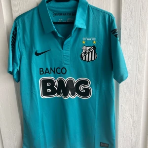 Santos FC Neymar Jr fotbollströja - Säljer en ljusblå Santos FC fotbollströja med Neymar Jr och nummer 11 på ryggen. Tröjan har svarta tryck, klubbmärke på bröstet och sponsorer på ärmar och framsida. Klassisk krage och kort ärm. Perfekt för fans av brasiliansk fotboll.