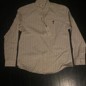 Vit randig skjorta från Ami Alexandre Mattiussi - Säljer en vit skjorta med svarta ränder från Ami Alexandre Mattiussi. Skjortan har lång ärm, klassisk krage och en röd broderad logga med hjärta och bokstaven A på bröstet. Aldrig använd, köpt på loppis (enligt säljaren). Mycket bra skick.