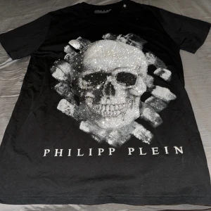 Svart t-shirt med dödskalle från Philipp Plein - Säljer en svart t-shirt från Philipp Plein med en stor glittrig dödskalle framtill och tydlig logga under motivet. T-shirten har korta ärmar och är i mjukt material. Perfekt för dig som gillar edgy och lyxig streetstyle. Betala genom Swish 