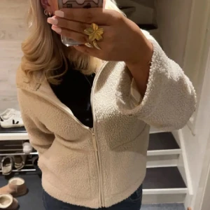 Beige teddyjacka med dragkedja - Mysig beige teddyjacka med dragkedja framtill. Perfekt för kyliga dagar med sin fluffiga och varma design. Jackan har en avslappnad passform och långa ärmar.