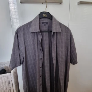 Grå kortärmad skjorta från Atlant - Säljer en grå kortärmad skjorta från Atlant med diskret rutigt mönster. Skjortan har klassisk krage och knäppning framtill. Perfekt för en stilren och avslappnad look.