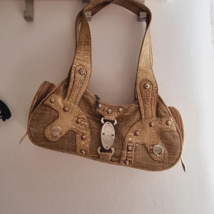 Guldig axelväska med metallicdetaljer - Säljer en unik guldig axelväska med croco-präglade detaljer och stora silvriga nitar. Väskan har dragkedja upptill och coola metalldetaljer framtill. Perfekt för dig som vill sticka ut med accessoarer!