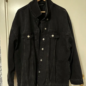 Svart jeansjacka - Säljer en baggy svart jeansjacka med klassisk design och silverfärgade knappar. Jackan har två bröstfickor med lock och knappar samt sidofickor. Passformen är oversized och materialet är kraftig denim. Perfekt för dig som vill ha en stilren och tidlös jacka.
