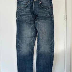 Blå jeans 32x32” - Klassiska blå Levis 501 jeans med raka ben och knappgylf. Jeansen har fem fickor och tydliga slitningar för en vintage look. Perfekta för dig som gillar tidlös denimstil.