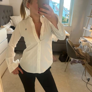 Vit skjorta från Polo Ralph Lauren - Klassisk vit skjorta från Polo Ralph Lauren med diskret broderad logga på bröstet. Skjortan har lång ärm, knappar framtill och en figurnära passform. Perfekt till både jeans och kostymbyxor. Storlek S🩷