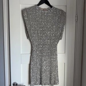 Glittrig silverklänning - Säljer en glittrig silverklänning med kort ärm och rund halsringning. Klänningen har en figurnära passform och är täckt av paljetter som ger ett riktigt festligt intryck.