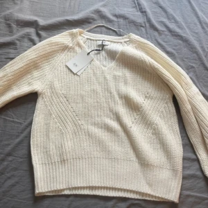 Vit stickad tröja från  - Säljer en vit stickad tröja från med v-ringning och lång ärm. Tröjan har ett strukturerat stickmönster och bred ribbad kant nedtill och vid ärmsluten. Perfekt för lager-på-lager och enkel att matcha.