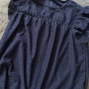 Marinblå blus med spetsdetaljer - Säljer en marinblå blus med korta ärmar och vackra spetsdetaljer samt broderad blomrad över bröstet. Klänningen har ett diskret hålmönster och rund halsringning. Perfekt för dig som gillar romantiska detaljer och en feminin stil.