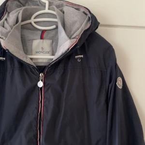 Mörkblå vindjacka från Moncler med huva - Snygg mörkblå vindjacka från Moncler med grå huva och dragkedja med röd, vit och blå detalj. Jackan har Moncler-logga på ärmen och är perfekt nu på våren/sommar. Väldigt bra skick och säljs då den blivit för liten. Jackan är i storlek 3 det vill säga small/medium.