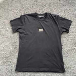 Svart t-shirt från Dolce & Gabbana - Säljer en svart t-shirt från Dolce & Gabbana med en metallbricka på bröstet där loggan står. T-shirten är i 100% bomull och har en klassisk passform med korta ärmar. Perfekt för dig som gillar stilrena och exklusiva plagg.