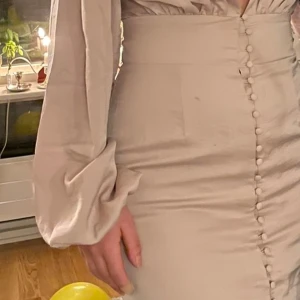 Beige omlottklänning från NA-KD - Säljer en beige omlottklänning från NA-KD med djup v-ringning och långa puffärmar. Klänningen har dekorativa knappar framtill och är tillverkad i ett mjukt, glansigt material som ger en elegant känsla.