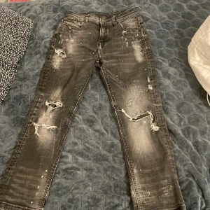 Svarta slitna jeans från Supply & Demand - Svarta jeans med slitningar och trasiga detaljer från Supply & Demand. Jeansen har en tvättad look med ljusa partier och raka ben. Klassisk femficksmodell och knappgylf. Perfekta för en edgy streetstil. Barn från 10-12 år storlek M 