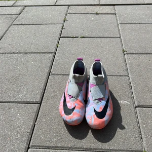 Nike Air Zoom fotbollsskor i grått och rosa - Nike Air Zoom fotbollsskor med grå ovandel och rosa samt ljusblått kamouflagemönster. Skorna har svart Nike-logga, elastisk stängning utan snörning och lila detaljer på hälen. Perfekta för dig som vill sticka ut på planen.