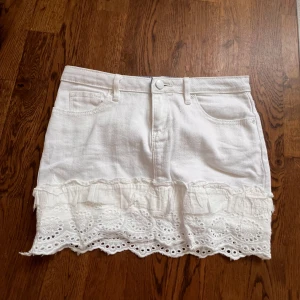 Vit jeanskjol med spetsdetaljer - Säljer en vit jeanskjol med volang och broderad spets längst ner. Kjolen har klassiska jeansfickor fram och bak samt knapp och dragkedja i midjan. Perfekt för dig som gillar romantiska detaljer och vill ha en söt look.💗