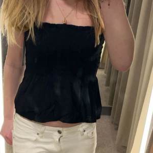 Svart topp med knytband - Säljer en svart topp med smockad överdel och volang nertill. Toppen har lite bredare axelband som knyts i rosetter på axlarna för en söt detalj. Använd 1-2 gånger endast!! Skriv om du har några frågor💕💕