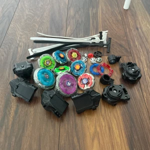 Beyblade burst set med snurror och avskjutare - Samling av flera färgglada Beyblade burst snurror i plast och metall, tillsammans med svarta avskjutare och vita ripcords. Perfekt för dig som gillar actionfyllda leksaksbattles. Innehåller olika modeller och tillbehör för att bygga egna kombinationer.
