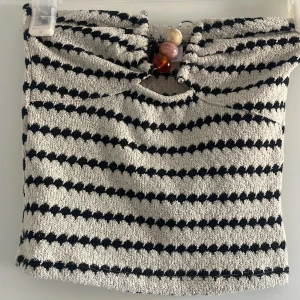 Randig tubtopp med pärldetalj - Säljer en beige och svart randig tubtopp med grovstickad känsla och dekorativa pärlor framtill. Toppen är croppad och har ett unikt mönster som ger en cool retrovibe. Perfekt för sommaren och festivaler!