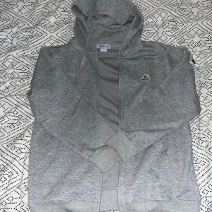 Grå hoodie från Moncler - Säljer en grå hoodie från Moncler med dragkedja framtill och huva. Tröjan har långa ärmar och en liten Moncler-logga på bröstet. Perfekt för en avslappnad stil.