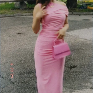 Rosa offshoulder långklänning - Säljer en snygg rosa långklänning med offshoulder-design och figurnära passform. Klänningen har draperade detaljer upptill och är perfekt för festliga tillfällen. Matchas snyggt med en rosa väska för en komplett look.