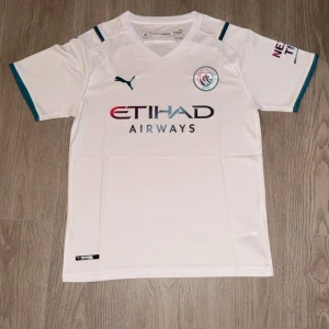 Manchester City vit bortatröja Puma - Säljer en vit Manchester City bortatröja från Puma med klubbmärke och sponsortryck på bröstet. Tröjan har korta ärmar och blå detaljer vid ärmslut. Perfekt för dig som vill visa ditt stöd för City på matchdag!