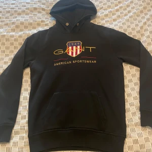 Svart hoodie från Gant - Svart hoodie från Gant med broderad logga och text på bröstet. Klassisk huva med dragsko och stor magficka framtill. Perfekt för en avslappnad och sportig stil.
