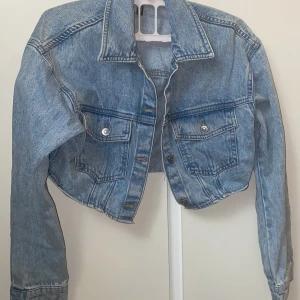 Croppad jeansjacka i ljusblå denim - Säljer en croppad jeansjacka i ljusblå denim med klassisk krage och två bröstfickor med knappar. Jackan har långa ärmar och silverfärgade knappar framtill. Perfekt för att ge en cool touch till din outfit. Den är XS  men passar nån som är M
