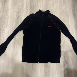 Svart zipjacka från BOSS med röda detaljer - Säljer en svart långärmad zipjacka från BOSS med röda detaljer längs axlarna och BOSS-logga på bröstet. Jackan har dragkedja hela vägen och en sportig look. Perfekt för dig som gillar stilrena plagg med diskreta detaljer.