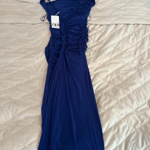 Marinblå långklänning från Zara - Säljer en marinblå långklänning från Zara med snygg rynkdetalj längs sidorna och axelband. Klänningen har en elegant, figurnära passform och är tillverkad i ett mjukt, stretchigt material. Perfekt för dig som vill sticka ut med stil.