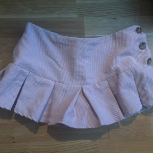 Ljusrosa manchesterkjol med knappar - Supersöt ljusrosa kjol i manchester med bred volang nertill och tre stora knappar i sidan. Kjolen har en kort och lekfull modell som passar perfekt till sneakers eller boots. Färgen är mer ljus rosa i verkligheten💗