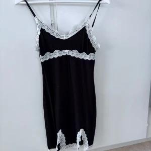 Svart slipklänning med spetsdetaljer - Svart slipklänning med tunna axelband och vita spetsdetaljer längs urringning, midja och nederkant. Klänningen har en feminin och elegant look, perfekt för dig som gillar en stilren men ändå söt stil.