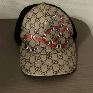 Gucci keps med ormtryck - Snygg keps från Gucci med klassiskt GG-mönster i beige och brunt samt en röd orm på framsidan. Baksidan är i svart mesh med justerbart spänne och röd-gröna detaljer. Perfekt för dig som vill sticka ut med en lyxig accessoar.