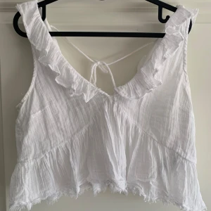 Vit volangprydd croppad linnetopp - Säljer en söt vit croppad linnetopp med volangdetaljer och v-ringning. Toppen har knytning i ryggen och är ärmlös, perfekt för varma dagar. Använd 2 gånger. Från Gina Tricot storlek S men stor i storleken. Jag har oftast M.