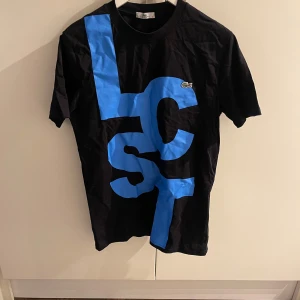 Svart t-shirt med blå tryck från Lacoste - Säljer en svart t-shirt från Lacoste med stort blått grafiskt tryck på framsidan och den klassiska krokodilloggan på bröstet. T-shirten har rund hals och korta ärmar. Perfekt för en cool och avslappnad stil.