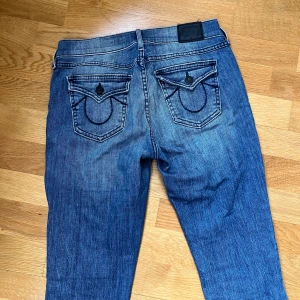Blå bootcut jeans från True Religion - Säljer ett par klassiska blå bootcut jeans från True Religion med snygga detaljerade bakfickor och kontrastsömmar. 