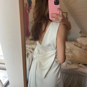 Vit klänning med rosett i ryggen - Säljer en elegant vit omlottklänning med djup v-ringning i ryggen och stor rosettdetalj bak. Klänningen är ärmlös och har en kort längd, perfekt för sommarens tillfällen. Materialet känns slätt och lätt.