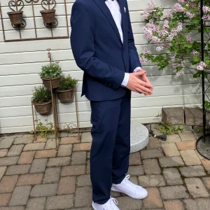 Mörkblå kostym från Dressmann - Säljers en stilren mörkblå kostym från Dressmann i slim fit-modell. Endast använd en gång! Kavajen har klassisk passform med två knappar, fickor med lock och bröstficka för näsduk. Skjortan ingår. Perfekt för bal eller student.                     Mått byxor: 71 cm från grenen till foten (insidan). 95 cm från midjan/bältet till foten (utsidan).  Mått kavaj: Ärm 59 cm, längd från kragen och ned 66 cm, bredd axlar 49 cm, bredd midja 40 cm. 